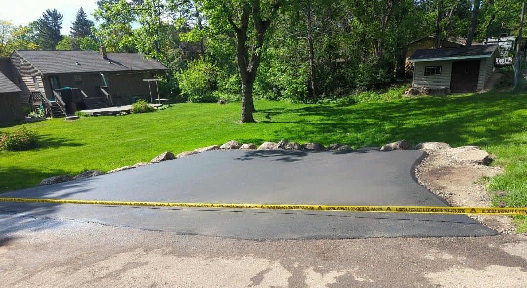 Freshly laid asphalt addition bordered by large rocks in a residential backyard in Becker, MN, marked off with yellow caution tape. 