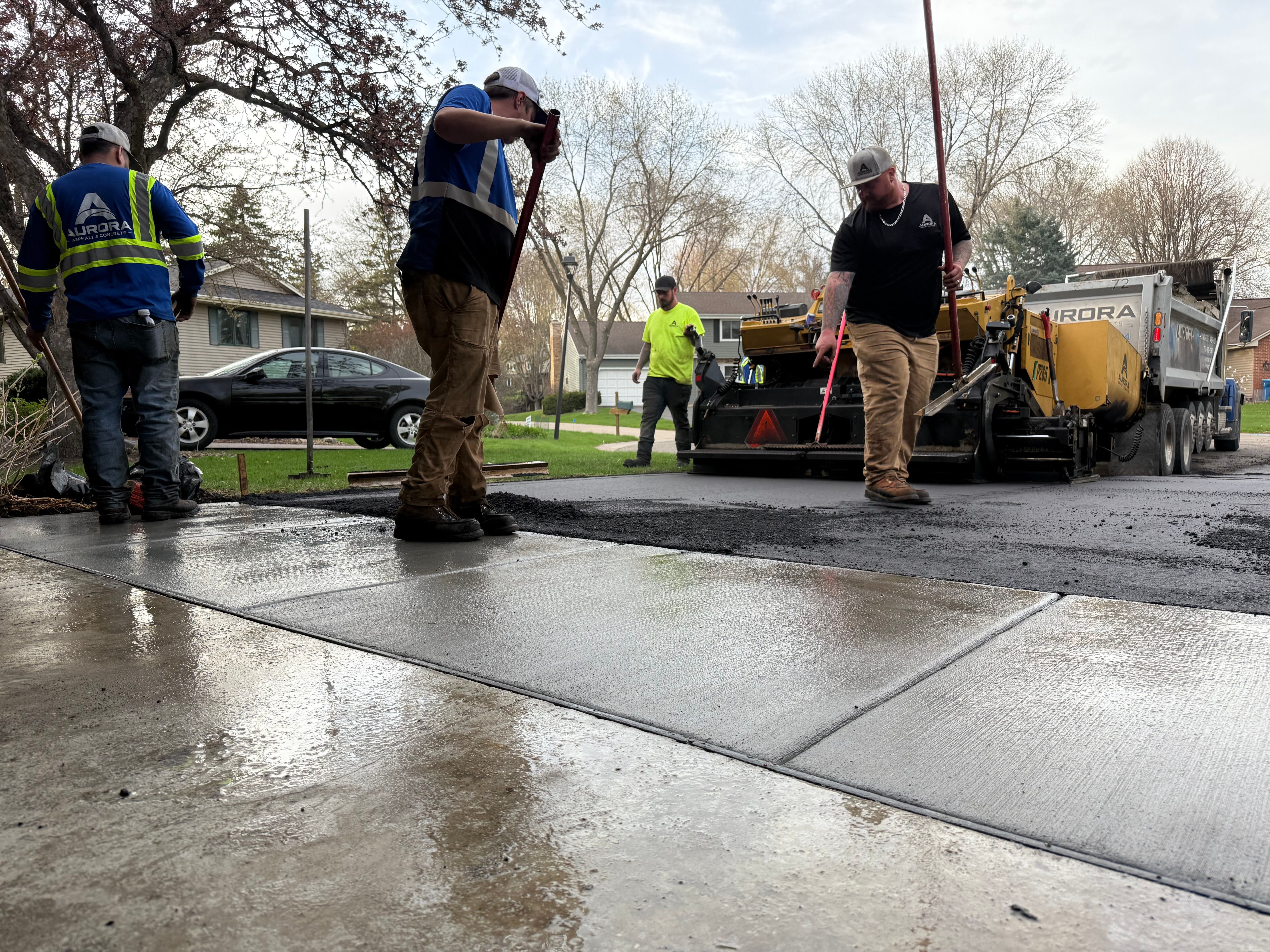 Construction crew spreading and leveling fresh asphalt during an asphalt driveway repair while a paving machine feeds new asphalt onto a residential driveway in Buffalo, MN.