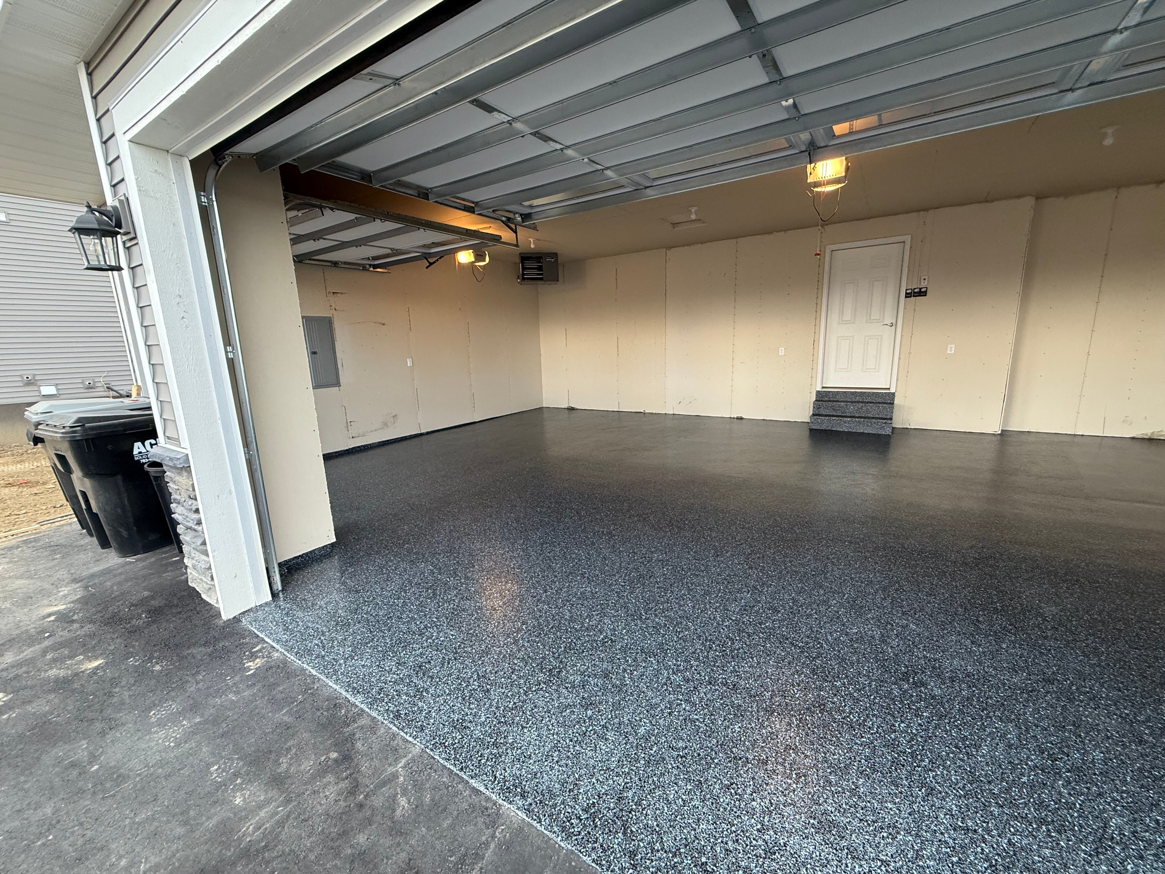 Clean, empty two-car garage with speckled epoxy-coated floor, beige unfinished drywall, and a white door with small steps leading into the house.