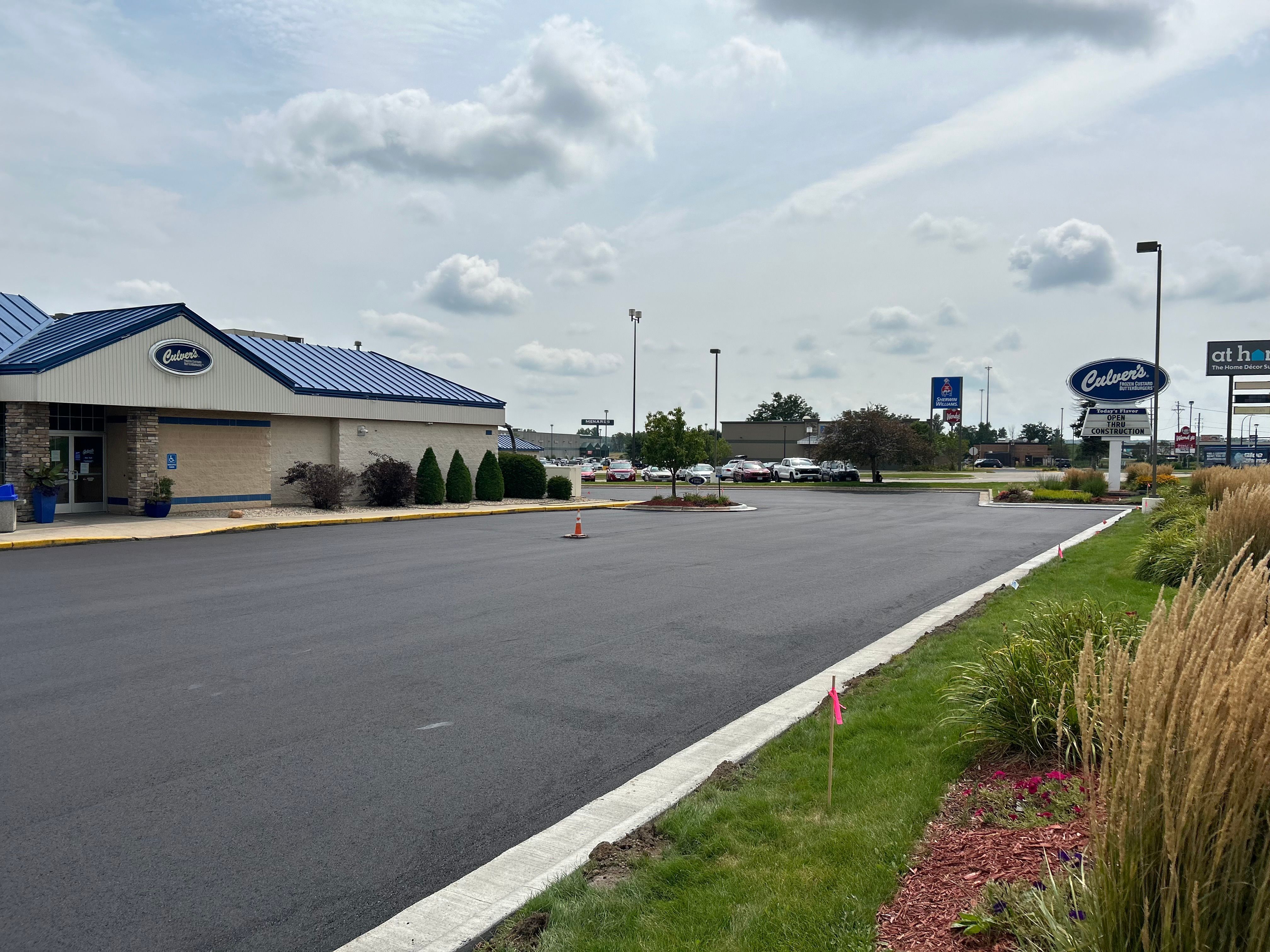 Freshly paved parking lot outside a Culver’s restaurant with a blue metal roof, surrounded by manicured landscaping and light cloud cover in the sky.