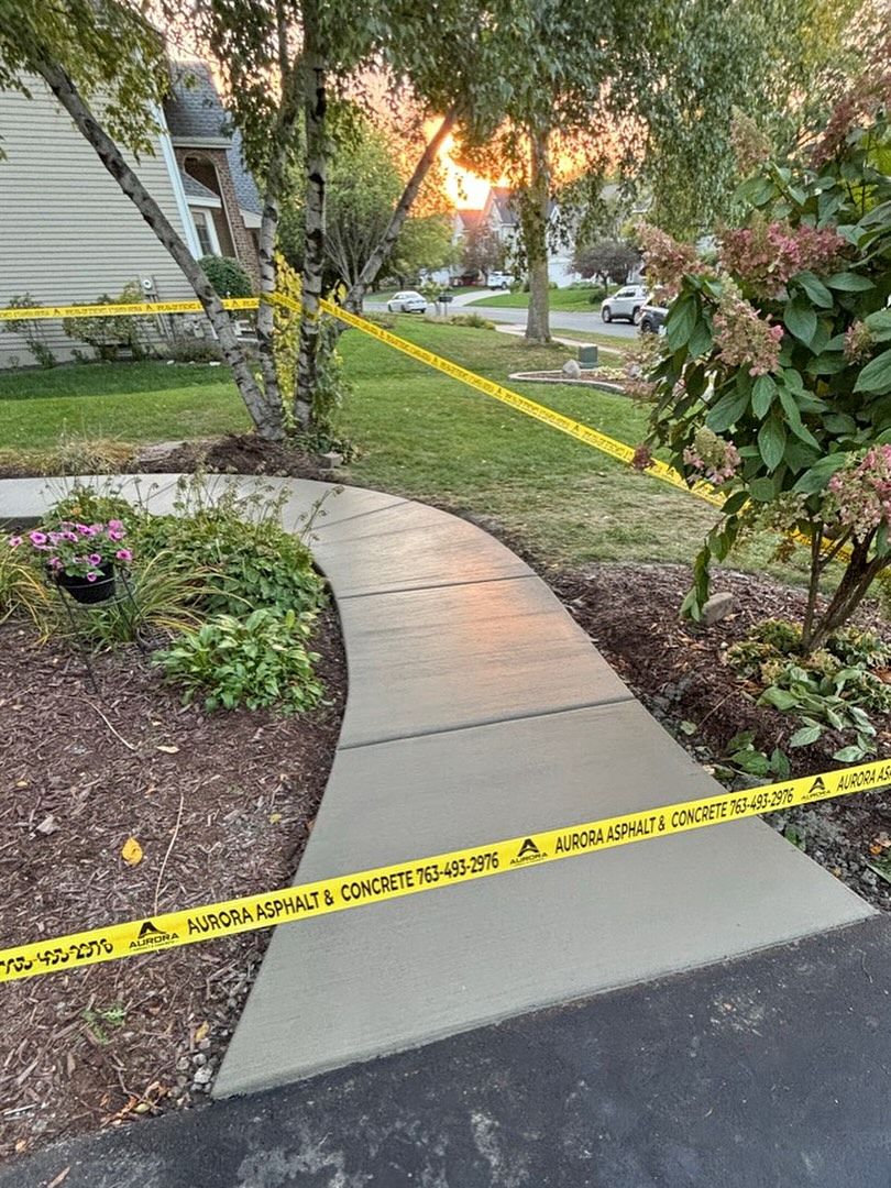 Freshly poured concrete sidewalk curves from a blacktop driveway through a landscaped front yard at sunset, bordered by mulch beds, flowers, and small trees in a Ramsey, MN neighborhood.